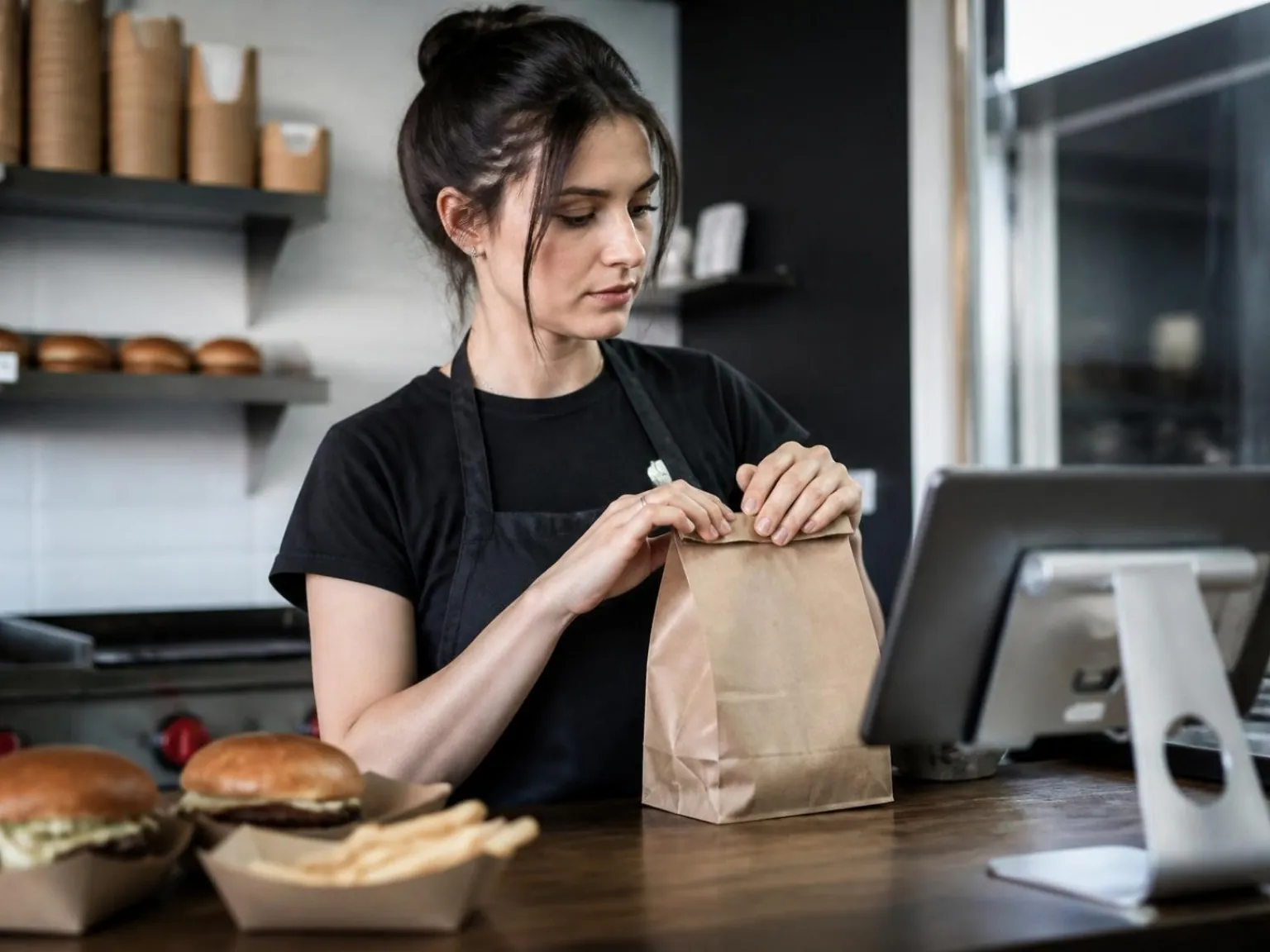 Employée qui ferme un sac de livraison au comptoir d’un restaurant de burgers, à côté d’une tablette de commande