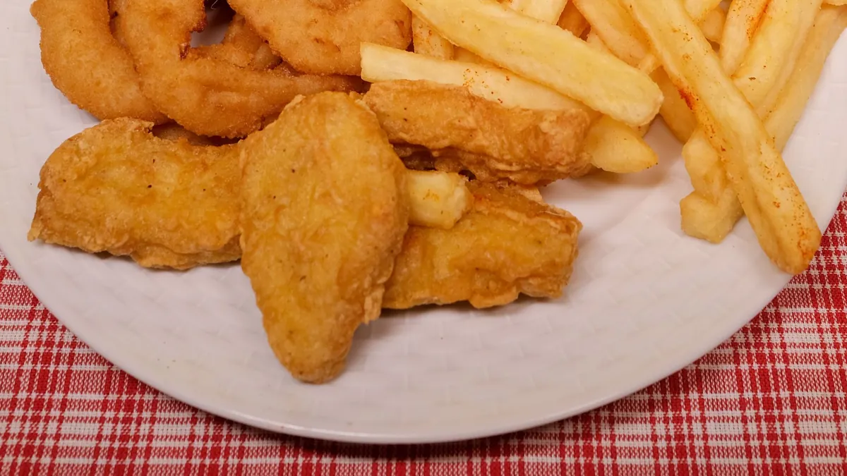 Assiette de morceaux de poulet pané et frit, avec des rondelles d’oignon et des frites, sur une nappe à carreaux rouges.