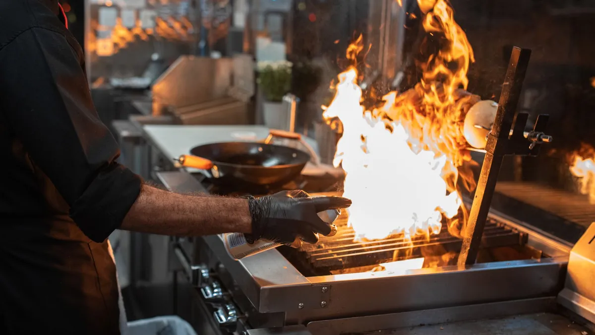 Un cuisinier ganté devant un grill en flammes, dans une cuisine professionnelle en inox.